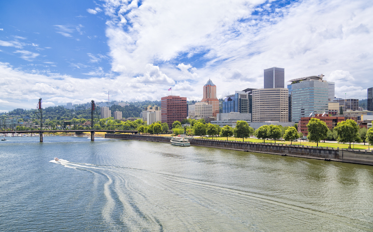 A motorboat leaves a gentle wake on the Willamette River as it passes the tree-lined Tom McCall Waterfront Park and the sun-drenched skyscrapers of downtown Portland.
