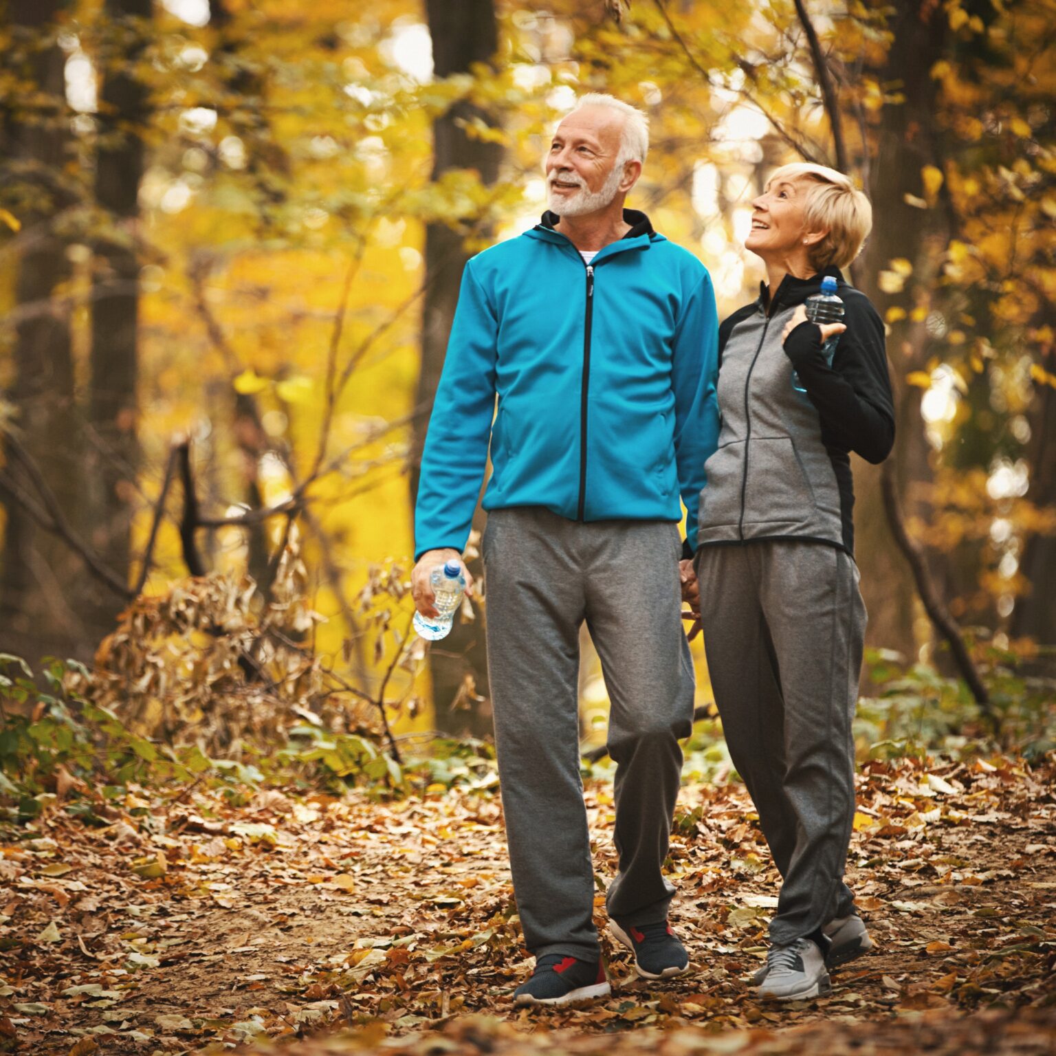 Older couple wearing windbreakers walking in the woods during fall, enjoying the beautiful foliage.